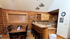 A cozy cabin-style kitchen with wooden walls, a dining table with chairs, a bar area, and a small trash bin. - Roomy Taylor Canyon Cabin With Private Fishing Access (Almont)
