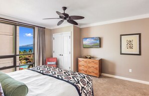 A hotel room with a bed, a ceiling fan, a view of the ocean, a door, a cabinet, and a framed picture on the wall.