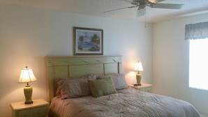 A bedroom with a bed, two bedside tables, a ceiling fan, a painting, and a window with curtains.