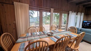 Una mesa de comedor de madera con seis sillas, un cuenco de madera y vistas a un lago y un bosque a través de amplios ventanales.