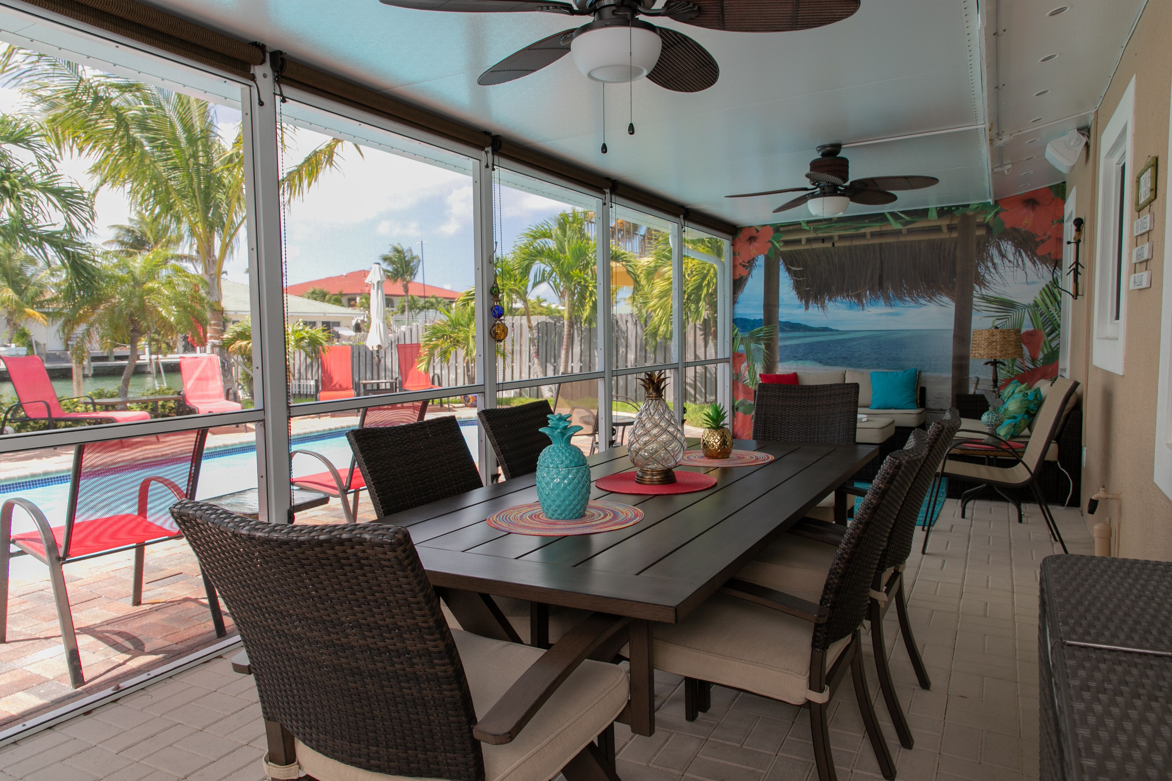 A sunlit dining area with a view of a pool and palm trees, featuring a long table with chairs and a ceiling fan.