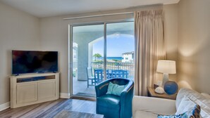 Ein Hotelzimmer mit Fernseher auf einem Schrank, einem Sofa, einem Sessel und einer Lampe, mit Blick auf den Strand.