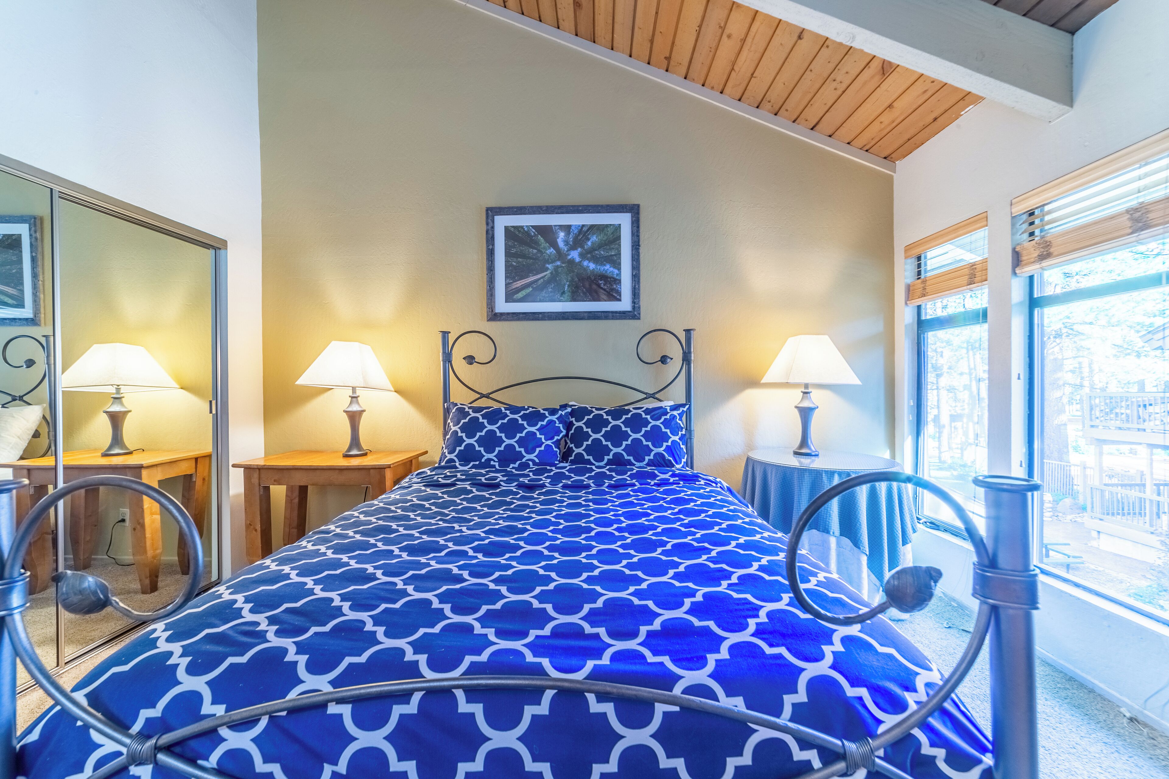 Een slaapkamer met een bed, twee nachtkastjes met lampen, een spiegel, een lijst met een afbeelding aan de muur en een raam met jaloezieën.