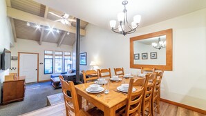 A dining area with a wooden table set for six, surrounded by wooden chairs. There is a chandelier above the table, a large mirror on the wall, and a door leading to another room.