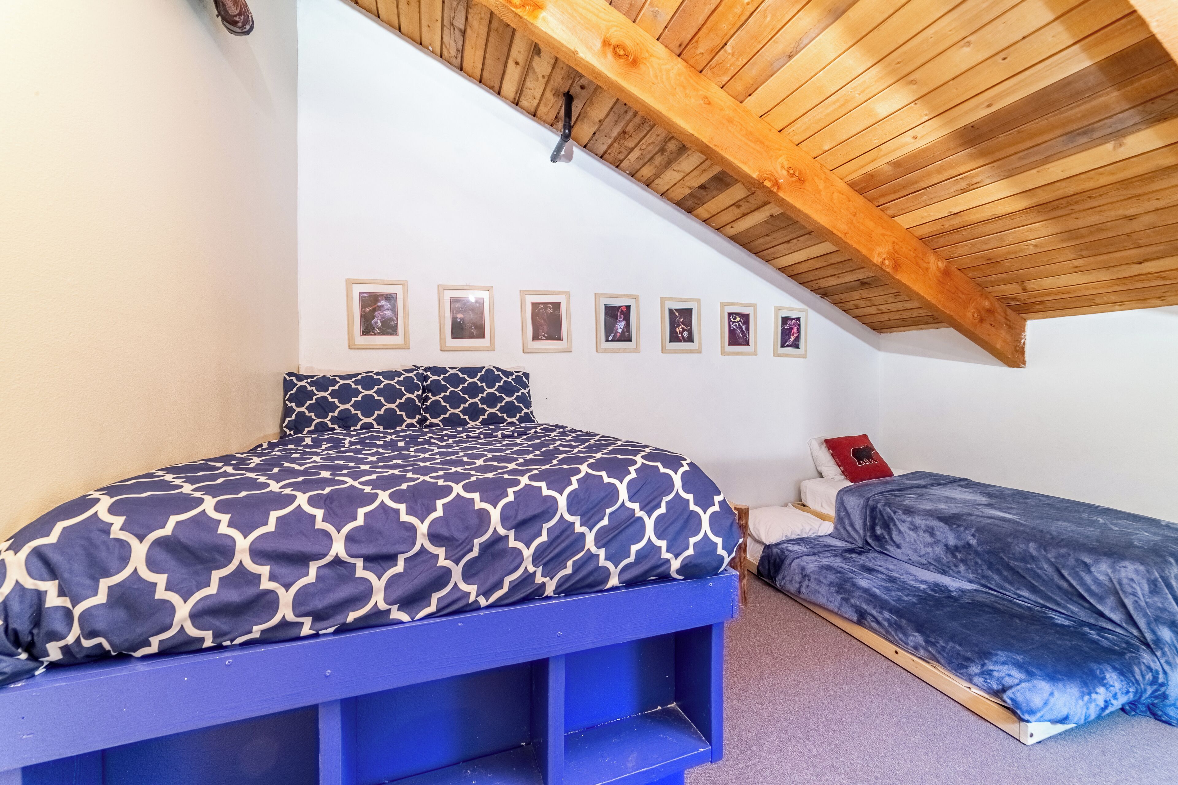 A bedroom with a sloped ceiling, a bed with a patterned bedspread, a smaller bed, and a series of framed pictures on the wall.