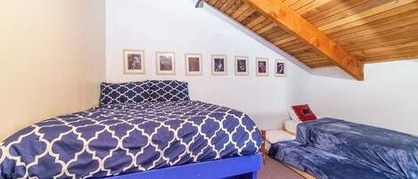 A bedroom with a sloped ceiling, a bed with a patterned bedspread, a smaller bed, and a series of framed pictures on the wall.