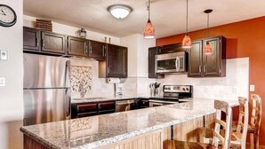 A modern kitchen with dark wooden cabinets, stainless steel appliances, a granite countertop, and wooden chairs. - Cimarron 301: Downtown Peak 9 Condo, Wood Fireplace, Pool & Sauna. (Breckenridge)