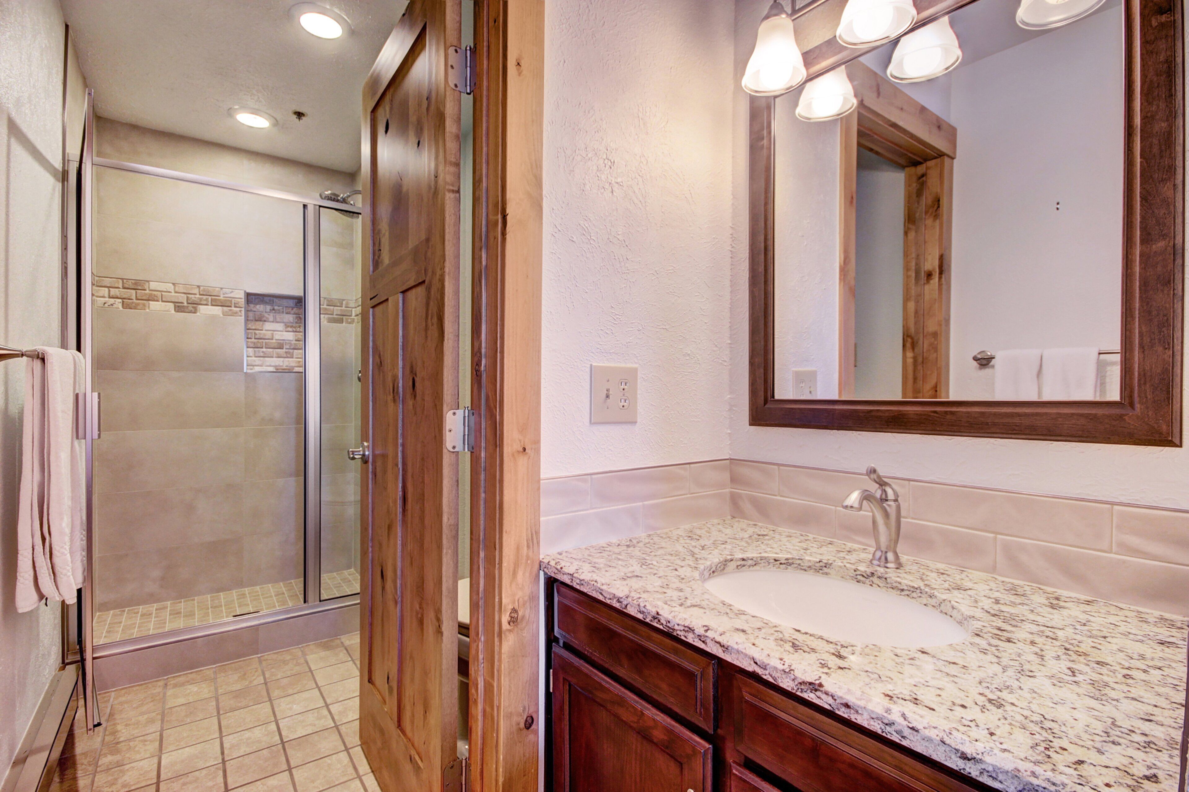 A bathroom with a glass shower door, a sink with a marble countertop, wooden cabinets, and a large mirror with lights above it.