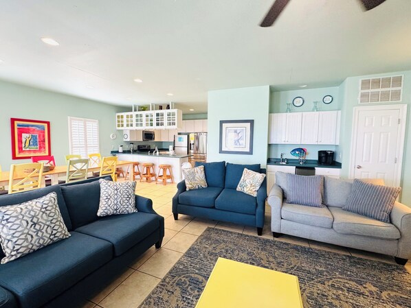A living room with blue sofas, a yellow coffee table, and a kitchen in the background.