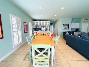 A dining area with a long table and colorful chairs, a kitchen in the background, and a living room with a sofa.