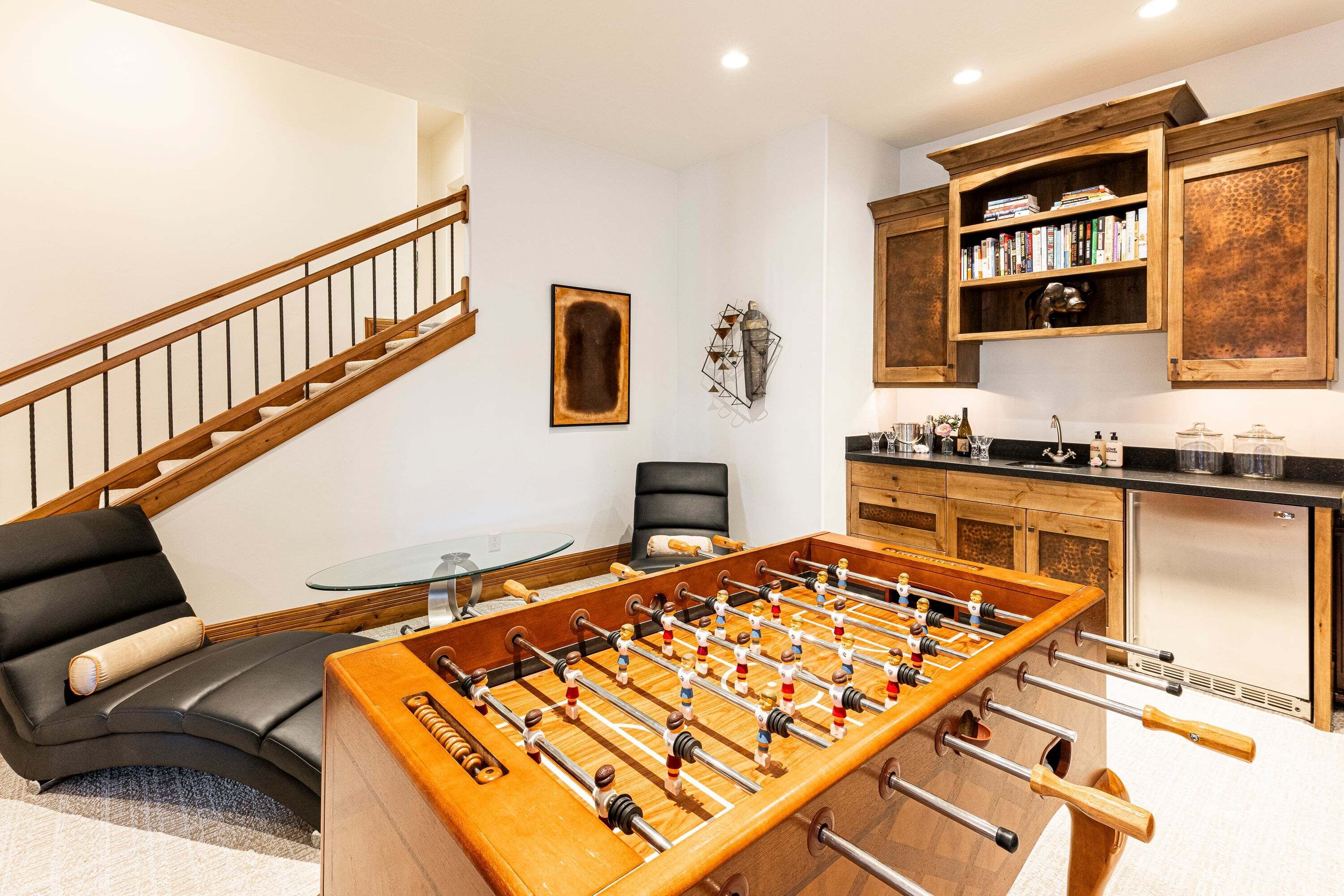 Un salon moderne comprenant une table de baby-foot, un canapé, un fauteuil, un escalier et un coin cuisine avec des meubles en bois et un réfrigérateur.