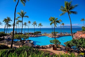 A resort pool area with multiple swimming pools, palm trees, and a view of the ocean.