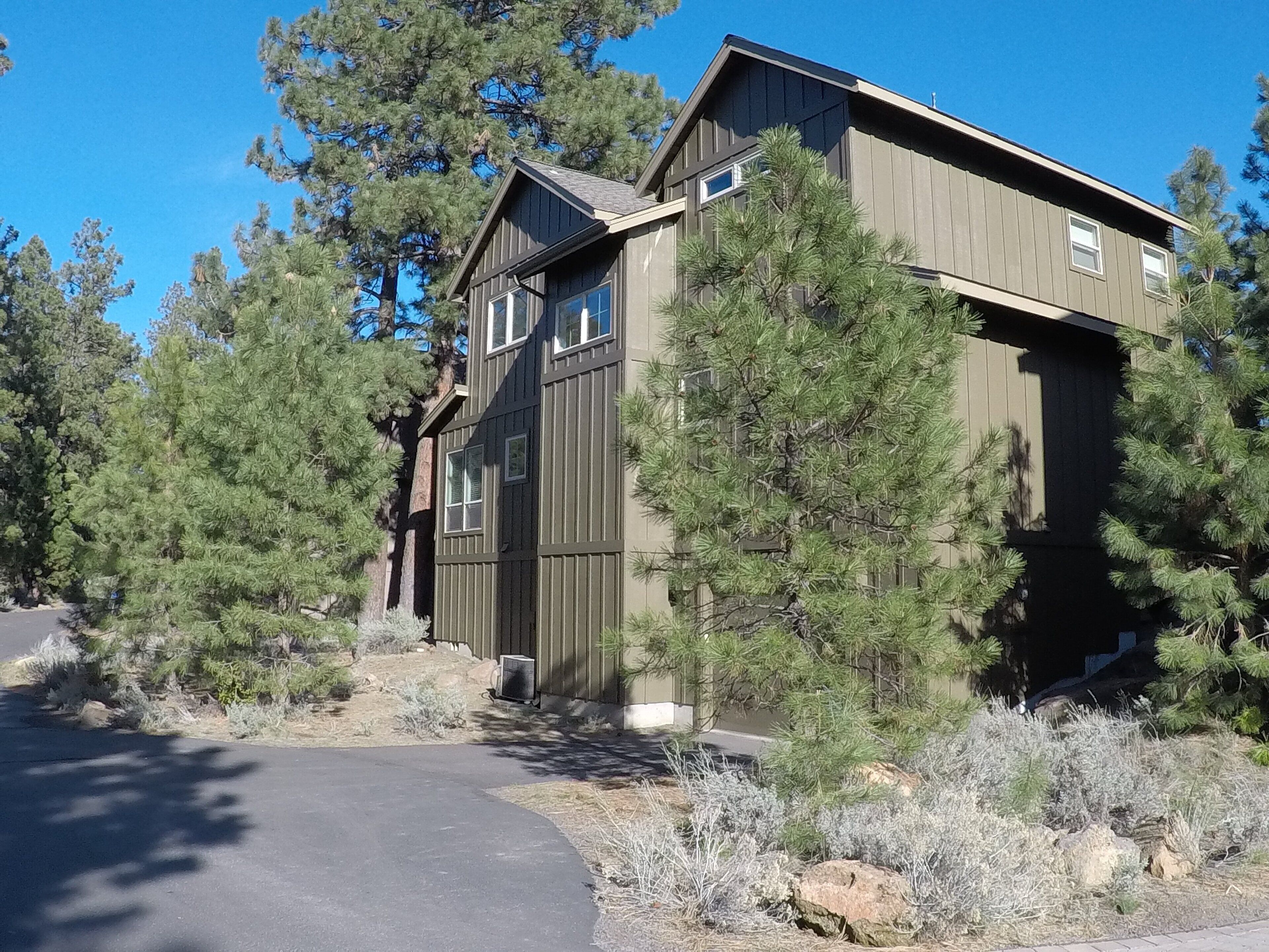 A two-story wooden cabin with a sloped roof, surrounded by pine trees and a paved driveway.
