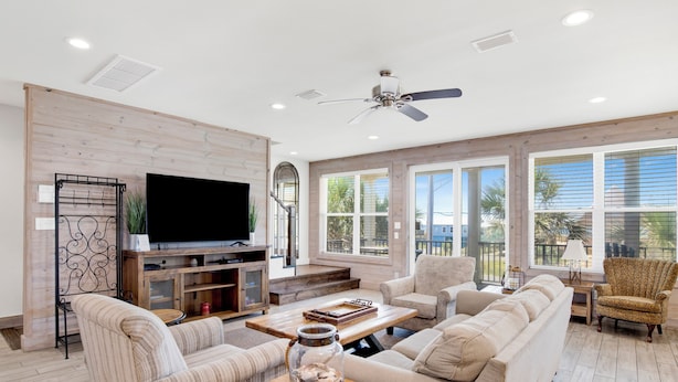 A spacious living room with a large flat-screen TV, a ceiling fan, and a wooden coffee table with a jar of seashells. - Pet Friendly | Community Pool | Beach Access (Gulf Shores)