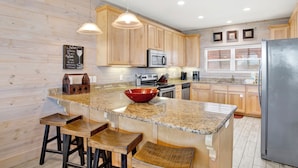 A kitchen with wooden cabinets, granite countertops, a large island with stools, a refrigerator, and a window with blinds. - Pet Friendly | Community Pool | Beach Access (Gulf Shores)