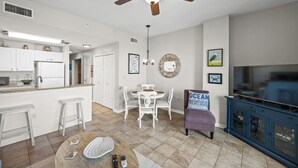 A modern kitchen with white cabinets, a dining area with a round table and chairs, a ceiling fan, and a TV cabinet.