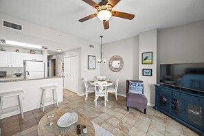 A modern kitchen with white cabinets, a dining area with a round table and chairs, a ceiling fan, and a TV cabinet.