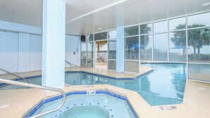 An indoor swimming pool with clear glass walls, a hot tub, and a view of palm trees outside.