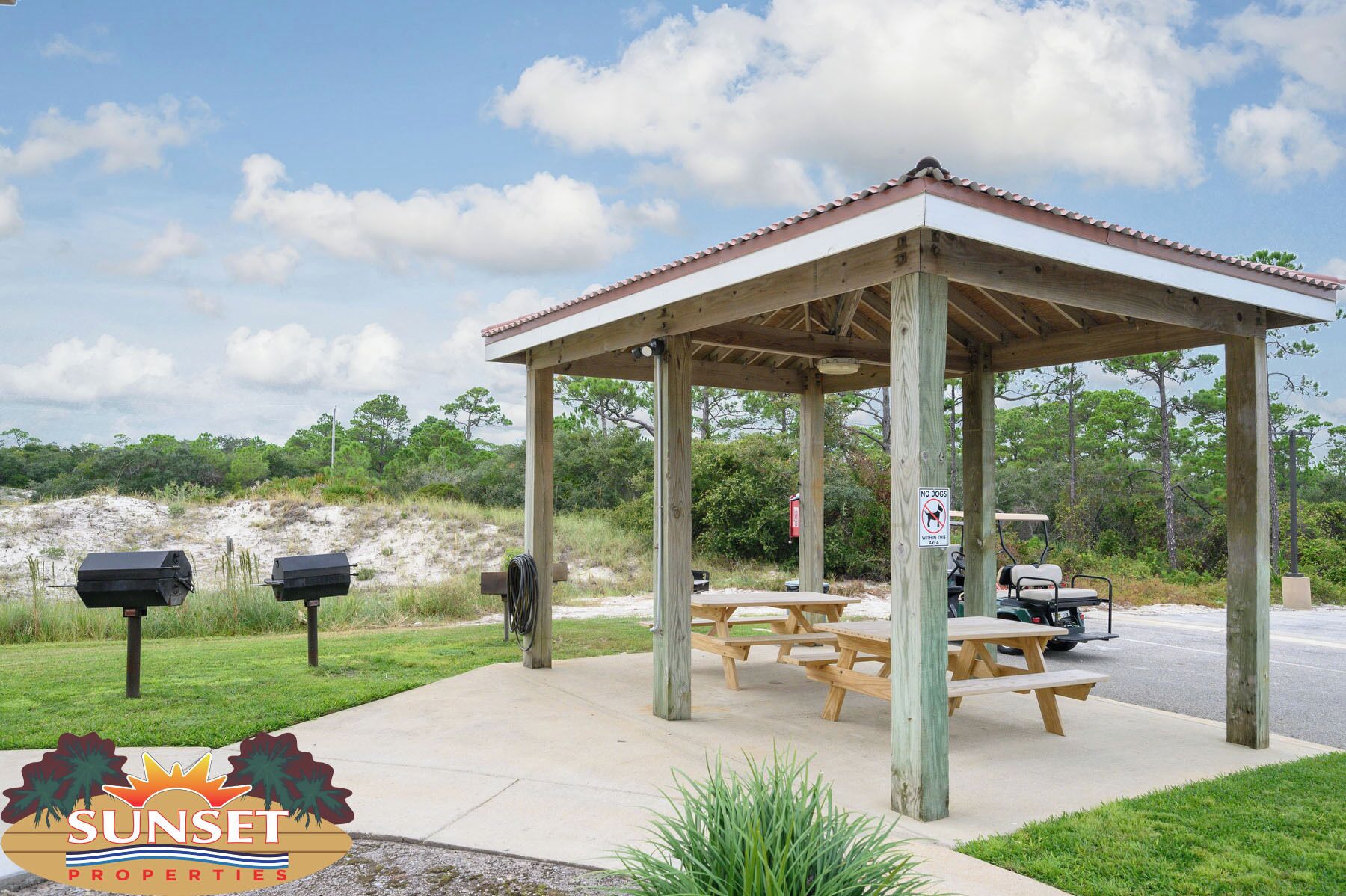 A wooden gazebo with picnic tables, surrounded by a grassy area and trees.