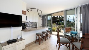 Una cocina moderna con un televisor grande, taburetes de bar y una mesa de comedor de vidrio con sillas.