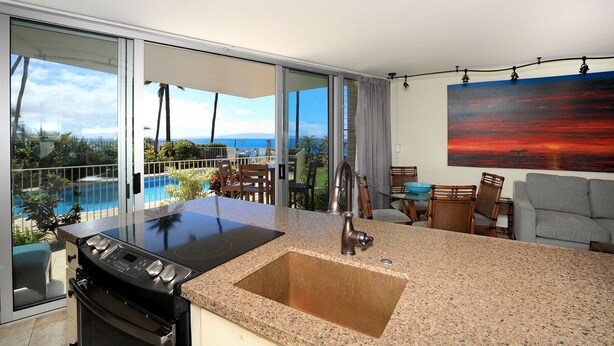 Cocina moderna con una isla grande, horno empotrado y un lavabo. Hay un comedor con sillas y una mesa, y un ventanal con vista a una piscina y al océano.
