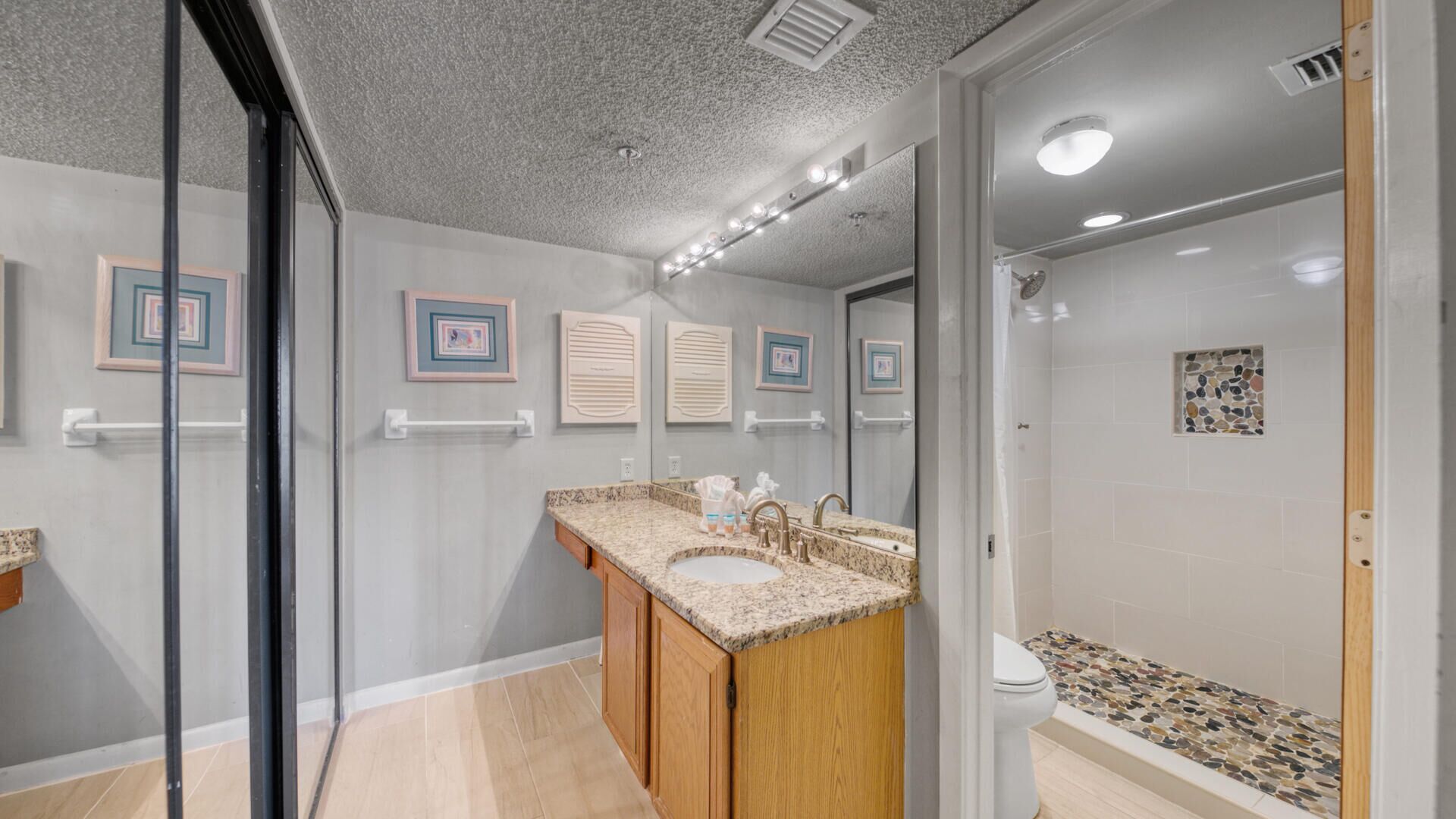 Une salle de bain moderne avec un lavabo surmonté d’un plan de travail en granit, des meubles en bois, une cabine de douche en verre et des œuvres d’art décoratives sur les murs.