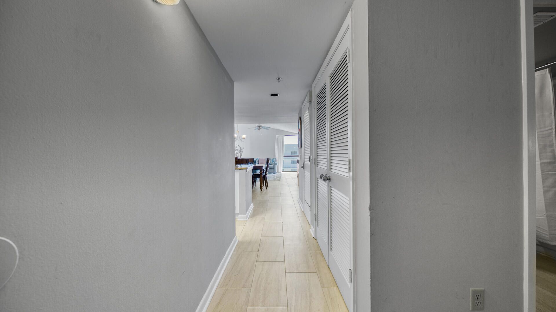 A narrow hallway with light-colored walls and wooden flooring, leading to a dining area with a table and chairs.