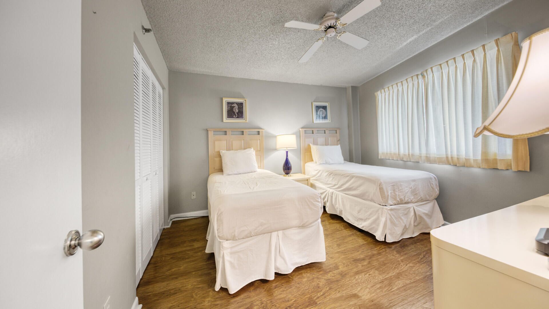A hotel room with two beds, a ceiling fan, a desk, and a window with curtains.