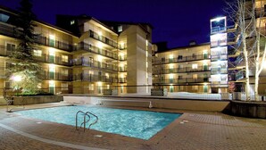 Piscina com deck revestido, rodeada por edifícios altos com sacadas e iluminação externa.