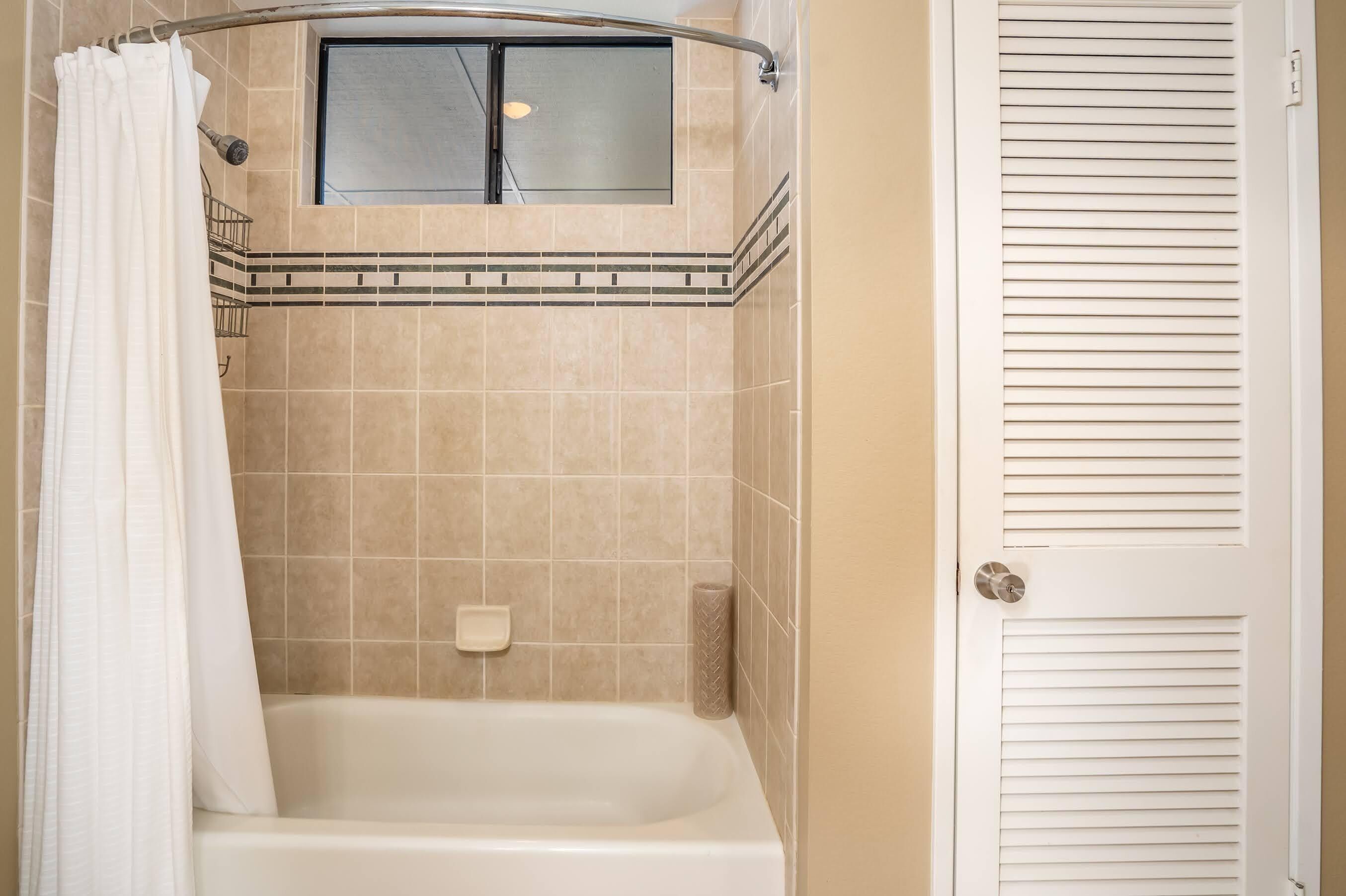 A bathroom with a bathtub, a shower curtain, a toilet paper holder, and a door with a louvered panel.