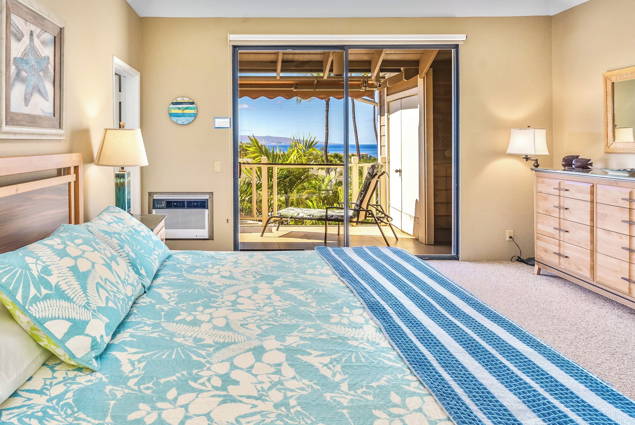 A bedroom with a bed, a dresser, a lamp, and a view of the ocean through an open door.