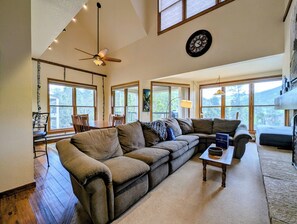 A spacious living room with a sectional sofa, a dining table with chairs, a ceiling fan, large windows, and a clock on the wall.
