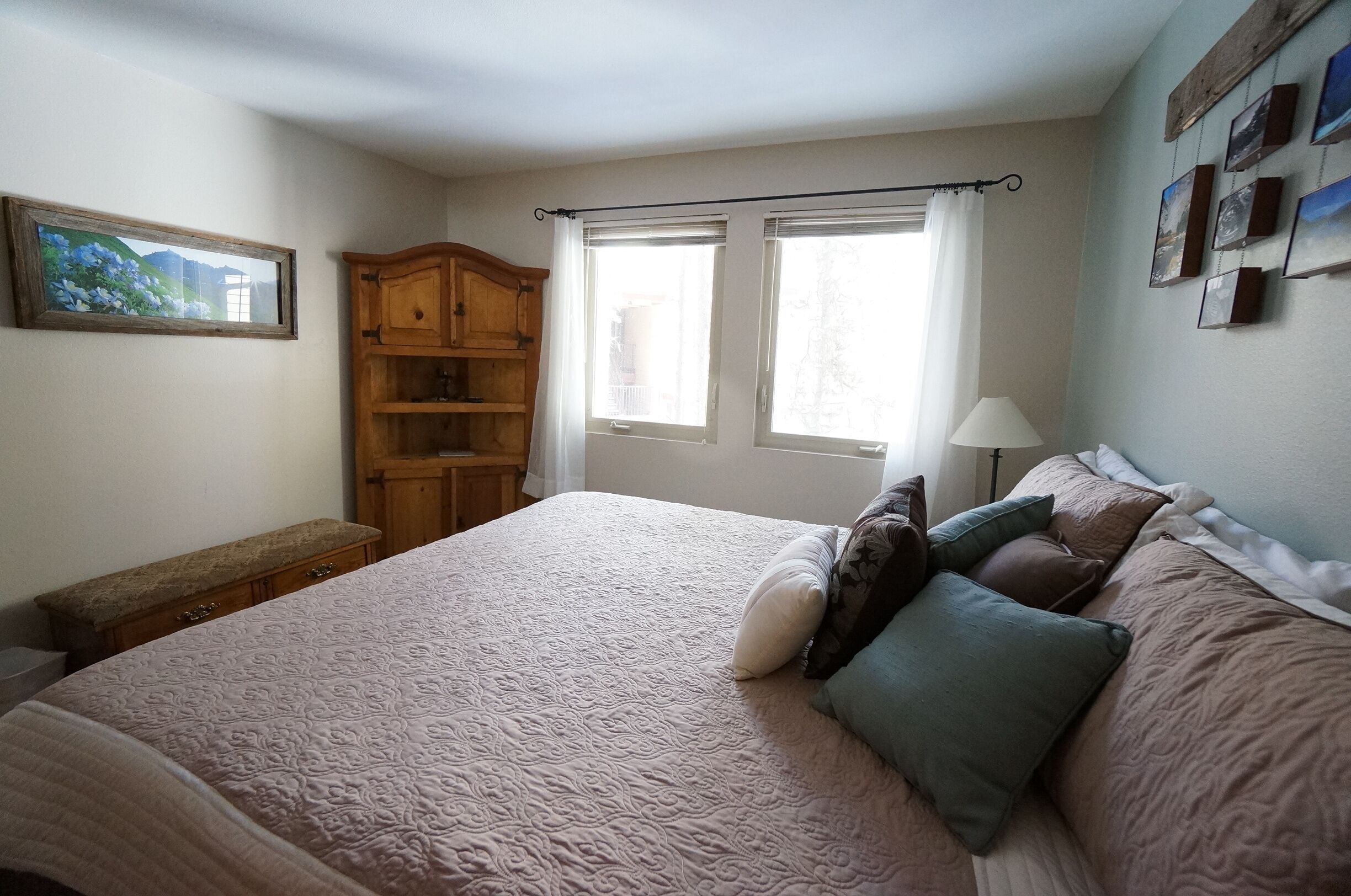 A bedroom with a large bed, a wooden wardrobe, two windows with curtains, and a wall-mounted picture frame.