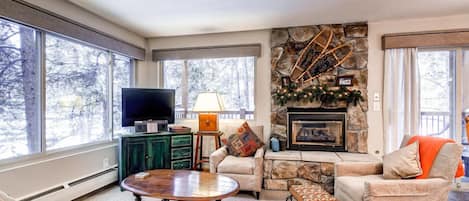A cozy living room with a stone fireplace, a television, and large windows.