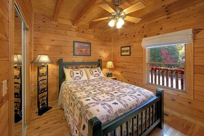 A cozy wooden cabin bedroom with a bed, a ceiling fan, a window with a view, and a wall-mounted lamp.