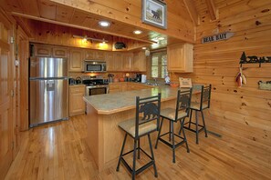 A wooden cabin interior with a kitchen featuring a refrigerator, microwave, stove, and wooden cabinets. There is a bar area with stools and a sign that reads "BEAR BAIT". - Cabin close to downtown w/Theater|Gameroom|Hot Tub (PIGEON FORGE)