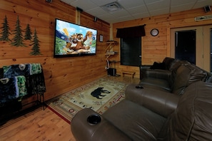 A cozy cabin interior with a wooden interior, a leather sofa, a television mounted on the wall, and a bear-themed rug. - Cabin close to downtown w/Theater|Gameroom|Hot Tub (PIGEON FORGE)