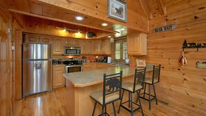 Interior de una cabaña de madera con cocina, que incluye refrigerador, microondas, estufa y armarios de madera. Cuenta con una barra con taburetes y un letrero que dice "BEAR BAIT".