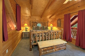 A log cabin bedroom with a wooden bed, two bedside lamps, a wooden bench, and a large window with curtains.