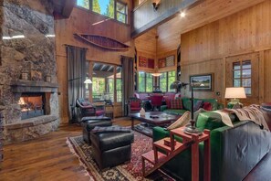 A spacious living room with a stone fireplace, wooden walls, and a large window overlooking a forest.
