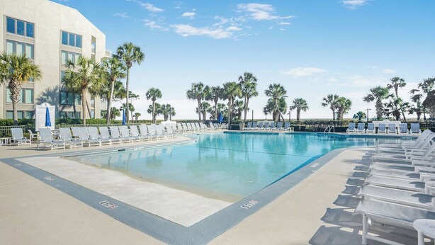 A pool area with lounge chairs and palm trees, surrounded by a building with multiple windows. - Shorewood 234-Freshly Updated in 2025 3BR Oceanview Near Shops & Restaurants (Hilton Head Island)