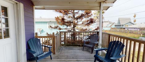 En veranda av trä med två Adirondack-stolar, en vit dörr och en utsikt över ett träd och hus i bakgrunden.