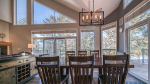 A spacious dining area with a large table and chairs, large windows offering views of trees, and a modern chandelier.