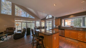 A spacious kitchen with a large island, wooden cabinets, and a view of trees through large windows.