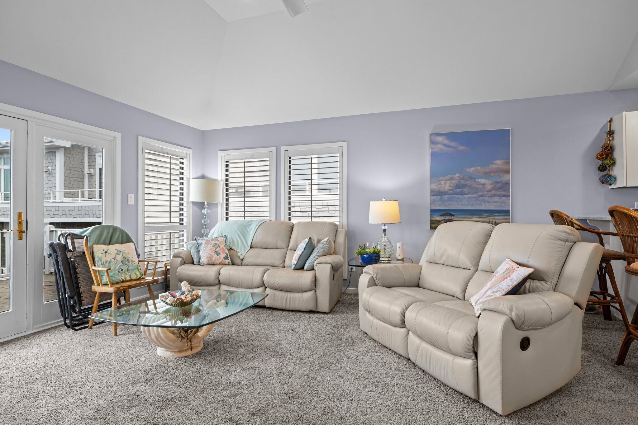 A living room with two beige sofas, a glass coffee table, and a view of the outside through large windows with shutters.