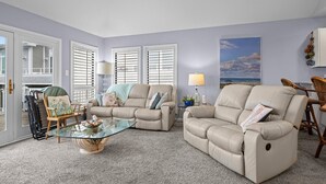 Ein Wohnzimmer mit zwei beigefarbenen Sofas, einem Glastisch und Blick ins Freie durch große Fenster mit Fensterläden.