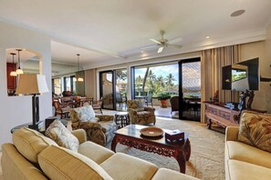 A spacious living area with a large sofa set, a coffee table, and a television. There is a balcony with a view of palm trees.