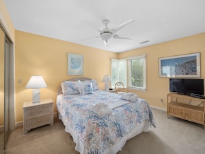 A bedroom with a bed, two bedside tables with lamps, a ceiling fan, a television, and a window with blinds.
