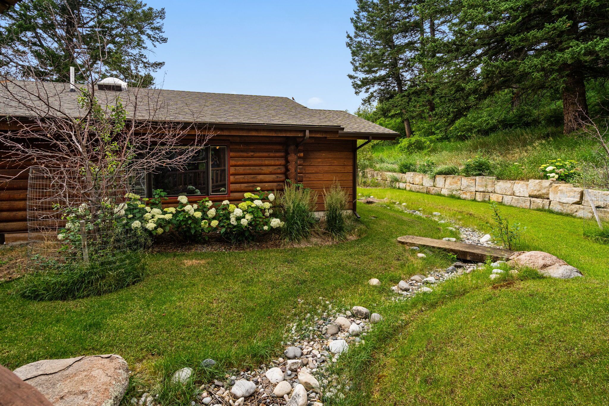 Una cabaña de troncos con un muro de contención de piedra, un jardín con césped y un pequeño arroyo.