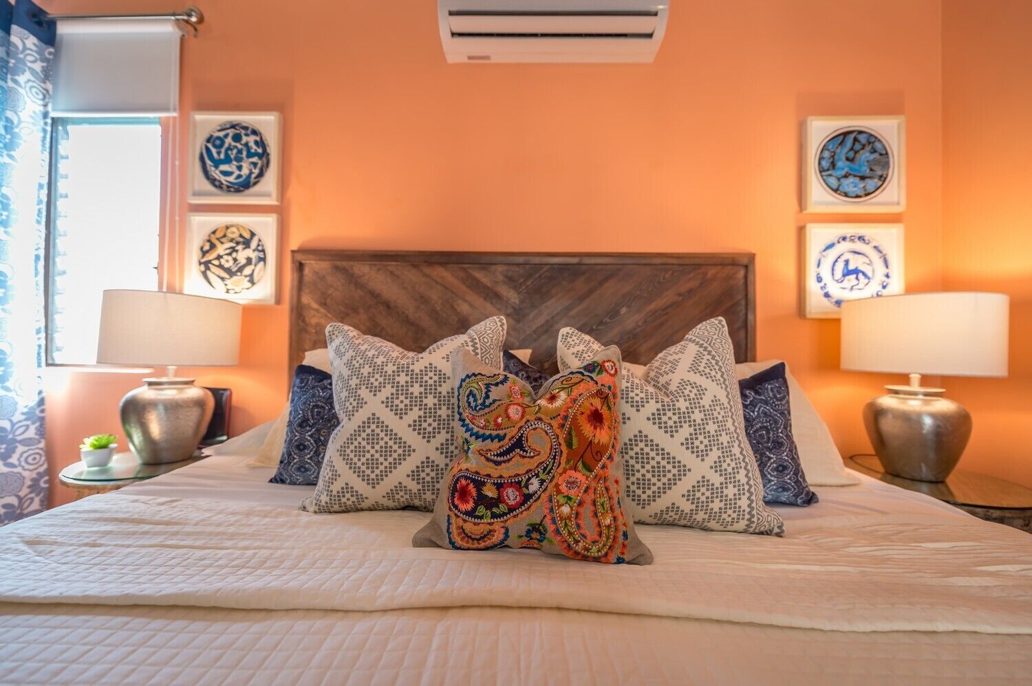 Une chambre d’hôtel avec un grand lit, des coussins décoratifs, des œuvres d’art mural et un climatiseur au plafond.